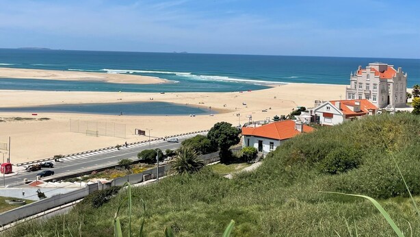 Paesaggio costiero con spiaggia, strada trafficata e un edificio dal tetto rosso.