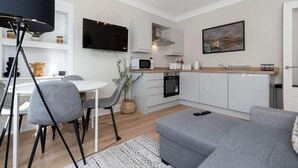 A modern kitchen with white cabinets, a microwave, and a sink. There is a dining area with a white table and grey chairs. A grey sofa is in the living area.