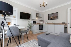 Eine moderne Küche mit weißen Schränken, einer Mikrowelle und einem Spülbecken. Es gibt einen Essbereich mit einem weißen Tisch und grauen Stühlen. Im Wohnbereich steht ein graues Sofa.
