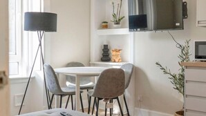 A dining area with a round table, four chairs, a lamp, a TV mounted on the wall, and a small shelf with decorative items.