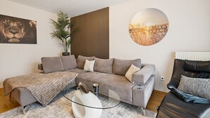 A modern living room with a grey sectional sofa, a glass coffee table, and a black leather armchair. There are two wall-mounted artworks, a potted plant, and a wooden panel wall. - Luxurious apartment I Fully equipped I University within walking distance (Homburg)