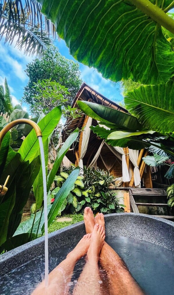 Una persona relajándose en un jacuzzi con exuberante follaje tropical y una estructura de madera al fondo.