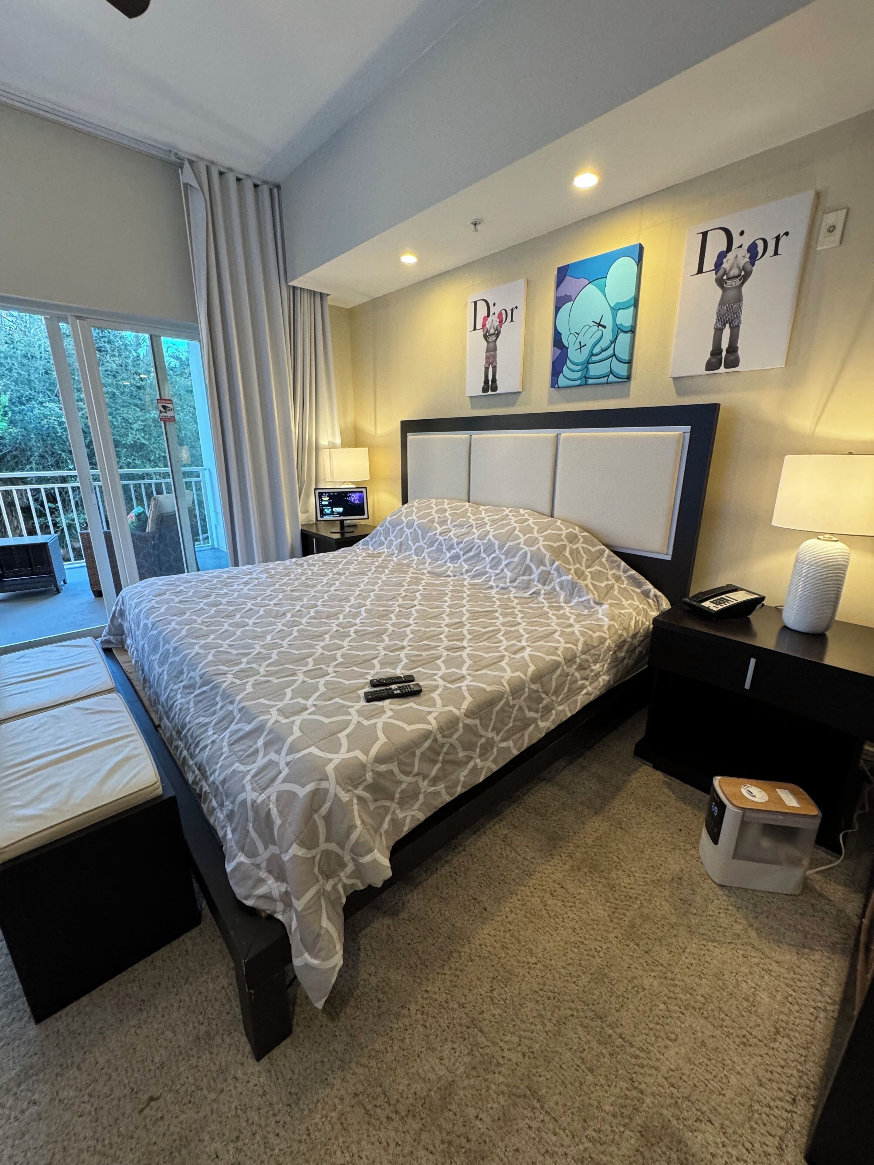 Una habitación de hotel con cama, mesita de noche, lámpara, cesto de basura y una obra de arte en la pared.