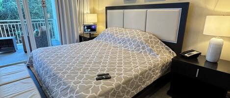 Una habitación de hotel con cama, mesita de noche, lámpara, cesto de basura y una obra de arte en la pared.