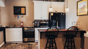 A kitchen with white cabinets, a black refrigerator, a microwave, and a coffee maker. There are two black bar stools at the island. - Forget your worries and enjoy some peace in the beautiful Lancaster Guest House. (New Holland)