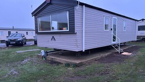 A mobile home with a large window, a door with a step, and a small porch area. - M&B Holiday,Cozy Beach side Caravan 4 Beds, 3 Sofa Bed, 7 Sleep,Wifi&Tea,Parking (Saint Osyth)