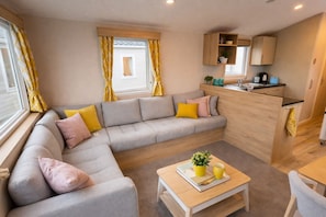 A modern living room with a grey sectional sofa, yellow cushions, a wooden coffee table, and a kitchen area with cabinets and a sink.