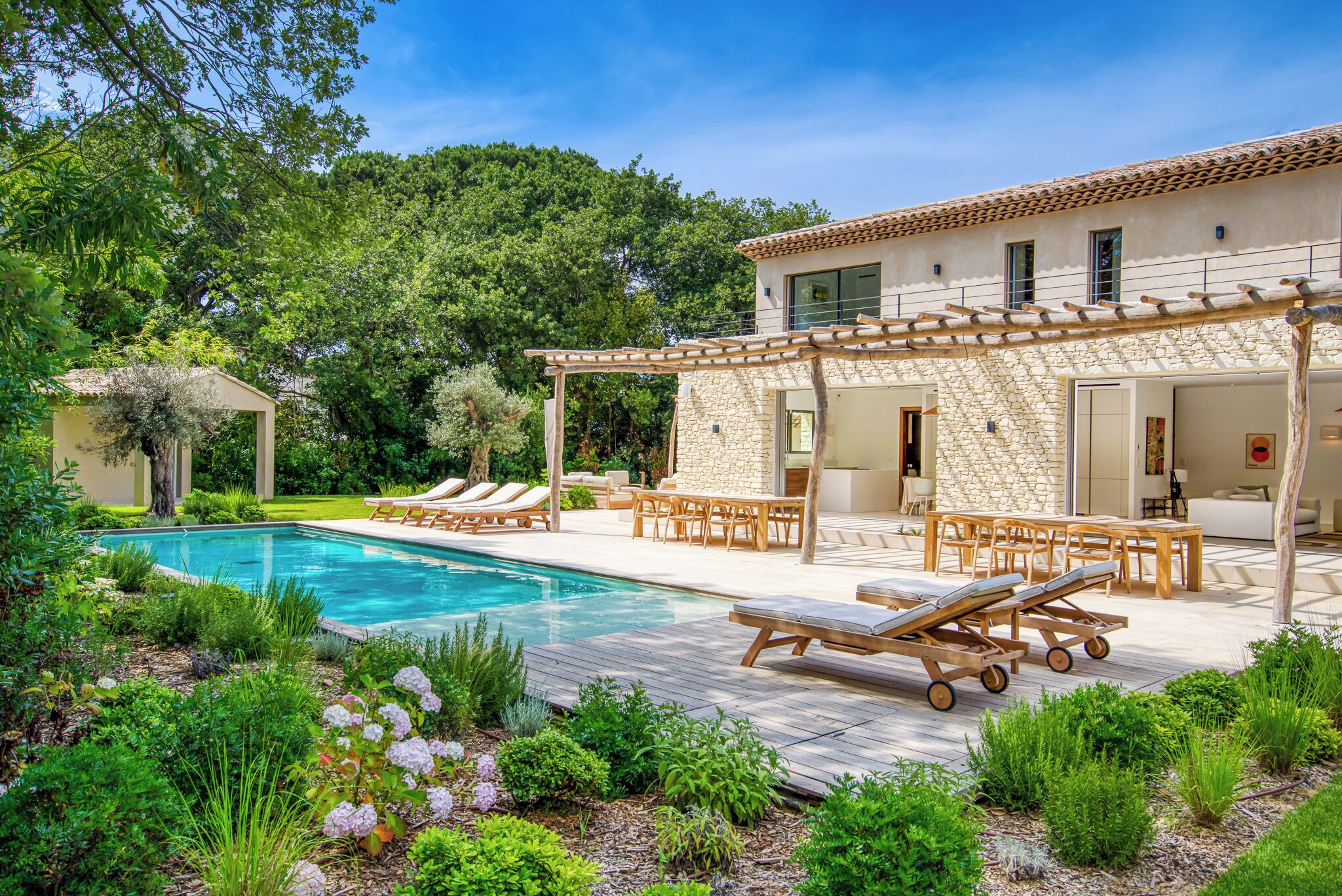 A poolside area with lounge chairs and a pergola, surrounded by lush greenery and a stone building.