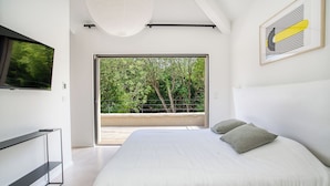 Ein modernes Schlafzimmer mit einem großen Bett, einem an der Wand montierten Fernseher und einem Balkon mit Blick auf Bäume.