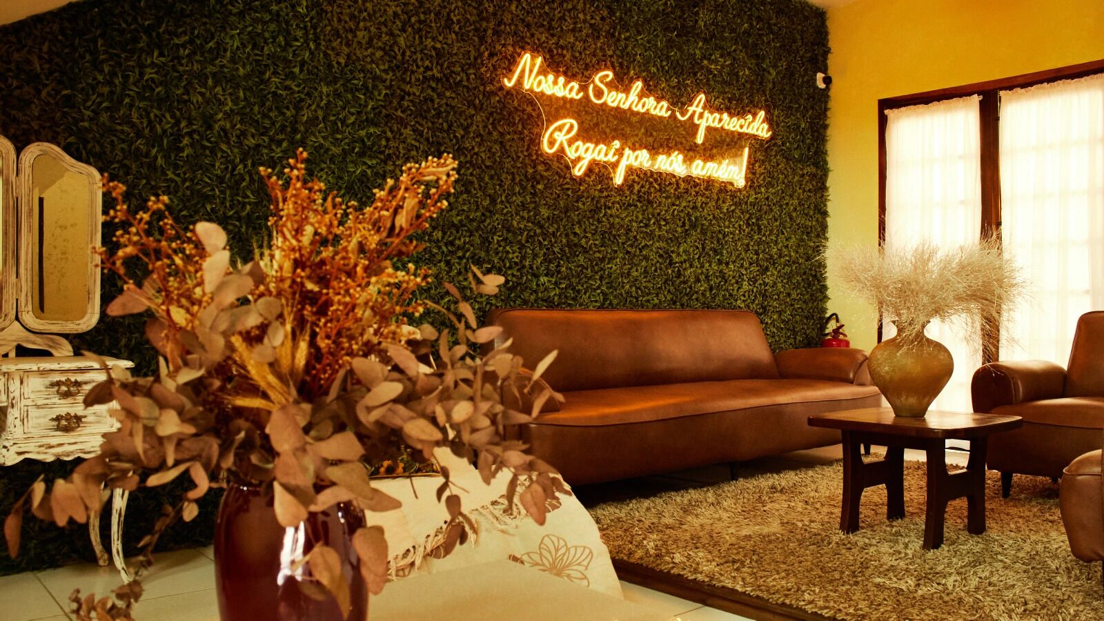 A cozy living room with a green wall, a neon sign, a brown leather sofa, and a vase with dried flowers.