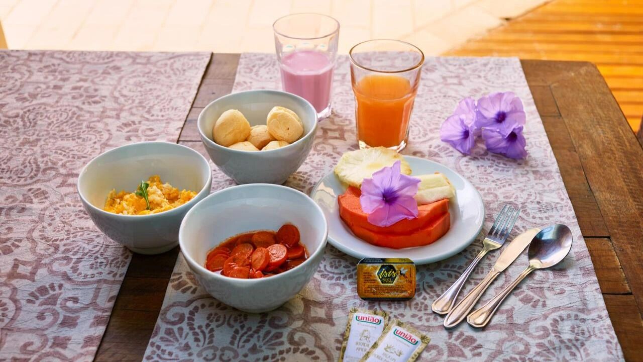 Una mesa con mantel estampado, dos platos de comida, un plato con una rebanada de fruta, un vaso de jugo de naranja y un pequeño paquete de té.