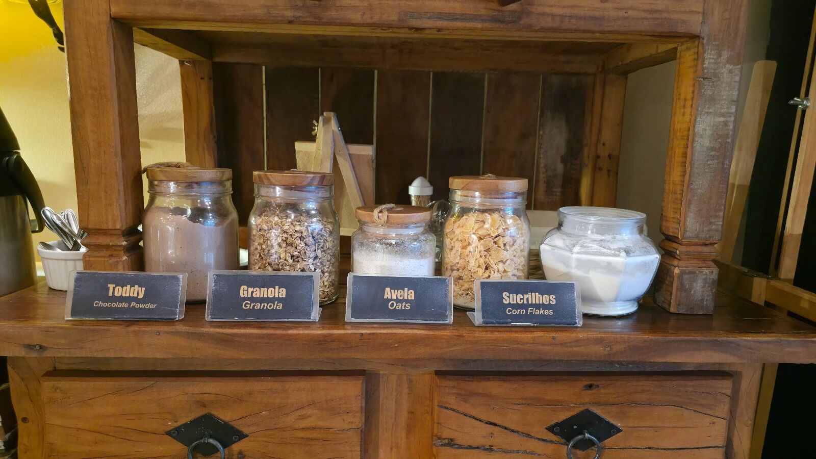 Un expositor de madera con frascos etiquetados que contienen granola, avena, cacao en polvo y copos de maíz.