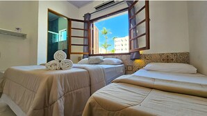 Quarto de hotel com duas camas, uma janela com cortinas, uma pilha de toalhas e vista para palmeiras e edifícios lá fora.