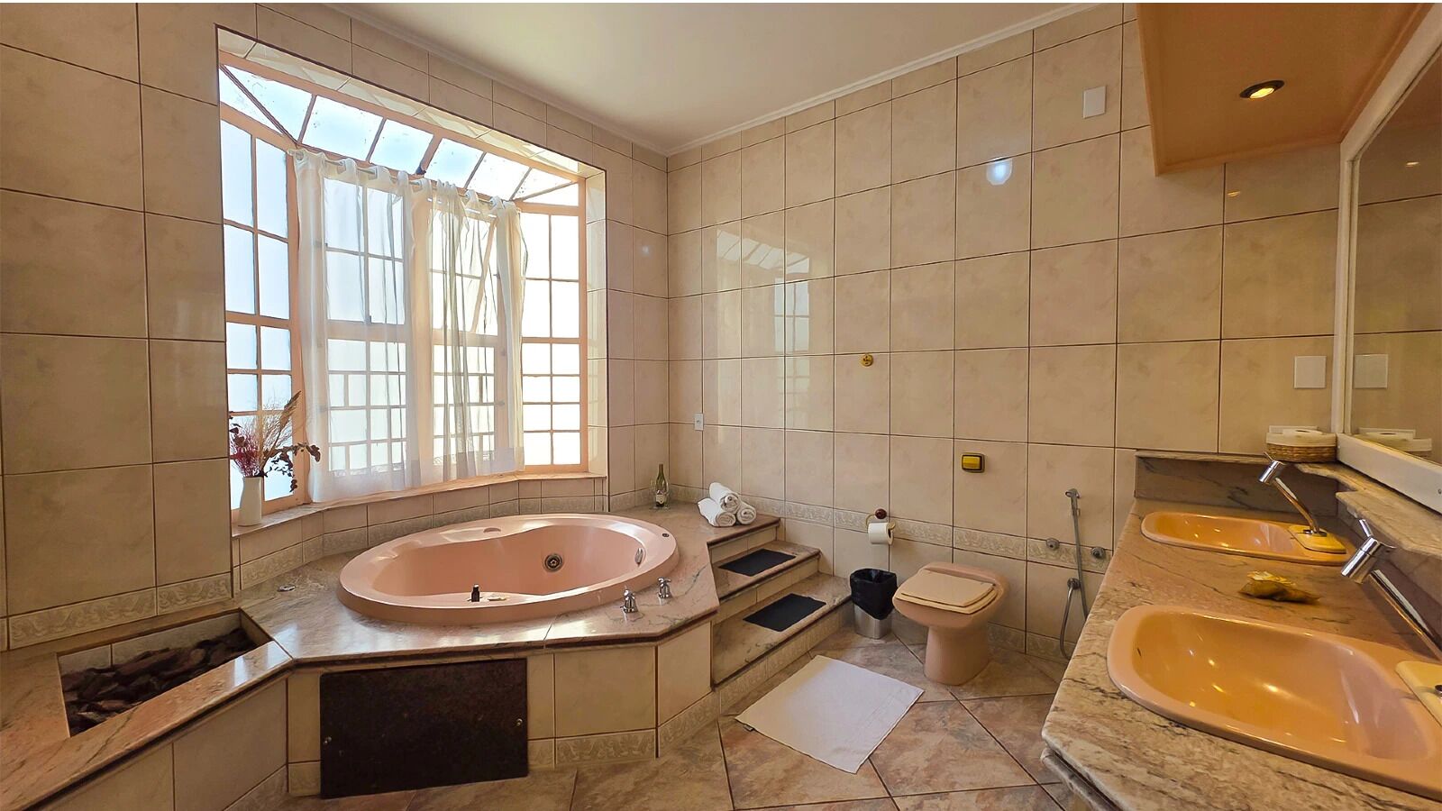A bathroom with a large oval sink, a toilet, and a bathtub. There is a window with curtains, a towel rack, and a trash bin.