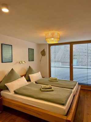 A double bed with green bedding, a wall-mounted light fixture, and a large window with blinds.
