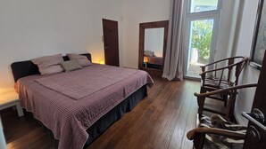 A bedroom with a large bed, wooden flooring, a window with curtains, and a wooden chair.