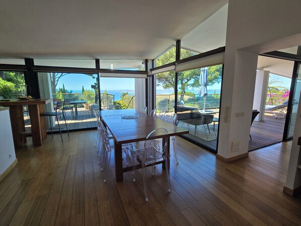 Une pièce spacieuse dotée d’un plancher en bois, de grandes fenêtres offrant une vue sur la mer, et d’un coin repas avec une table et des chaises.