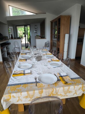 Un espace repas avec une longue table dressée pour le repas, des chaises transparentes et une vue sur l’extérieur.
