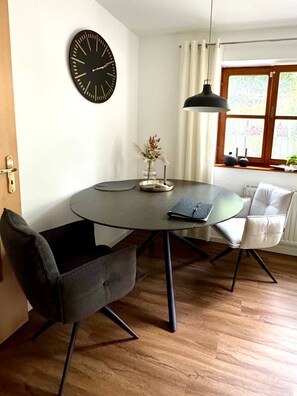 Ein Essbereich mit einem runden Tisch, zwei Stühlen, einer Uhr, einer Vase mit Trockenpflanzen und einem Fenster mit Vorhängen.