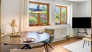 En matplats med ett runt bord, stolar, en tv och fönster med växter.