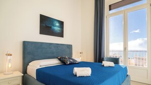 A neatly made bed with a blue bedspread and white pillows, a bedside table with a lamp, and a painting on the wall. - Residenza 2 Venti-Intera by Ortigia Apartments (Syracuse)