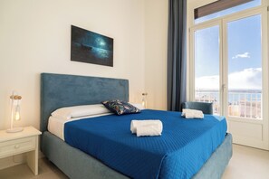 Ein ordentlich bezogenes Bett mit blauer Bettdecke und weißen Kissen, ein Nachttisch mit Lampe sowie ein Bild an der Wand.