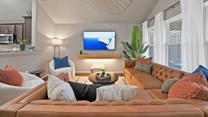 Ein modernes Wohnzimmer mit einem braunen Sofa, einem weißen Sessel und einem Couchtisch. Ein Flachbildfernseher ist über dem Sofa an der Wand montiert und zeigt eine schöne Landschaft.