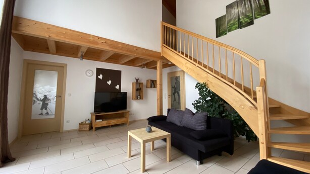 A living room with a wooden staircase, a black sofa, a TV, and a plant. - Holiday Home Plattenstein with Private Terrace, Garden, Sauna, Wi-Fi and BBQ (Kirchberg im Wald)