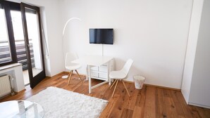 A modern room with a white desk, two white chairs, a mounted TV, a white rug, wooden flooring, and a door leading outside.