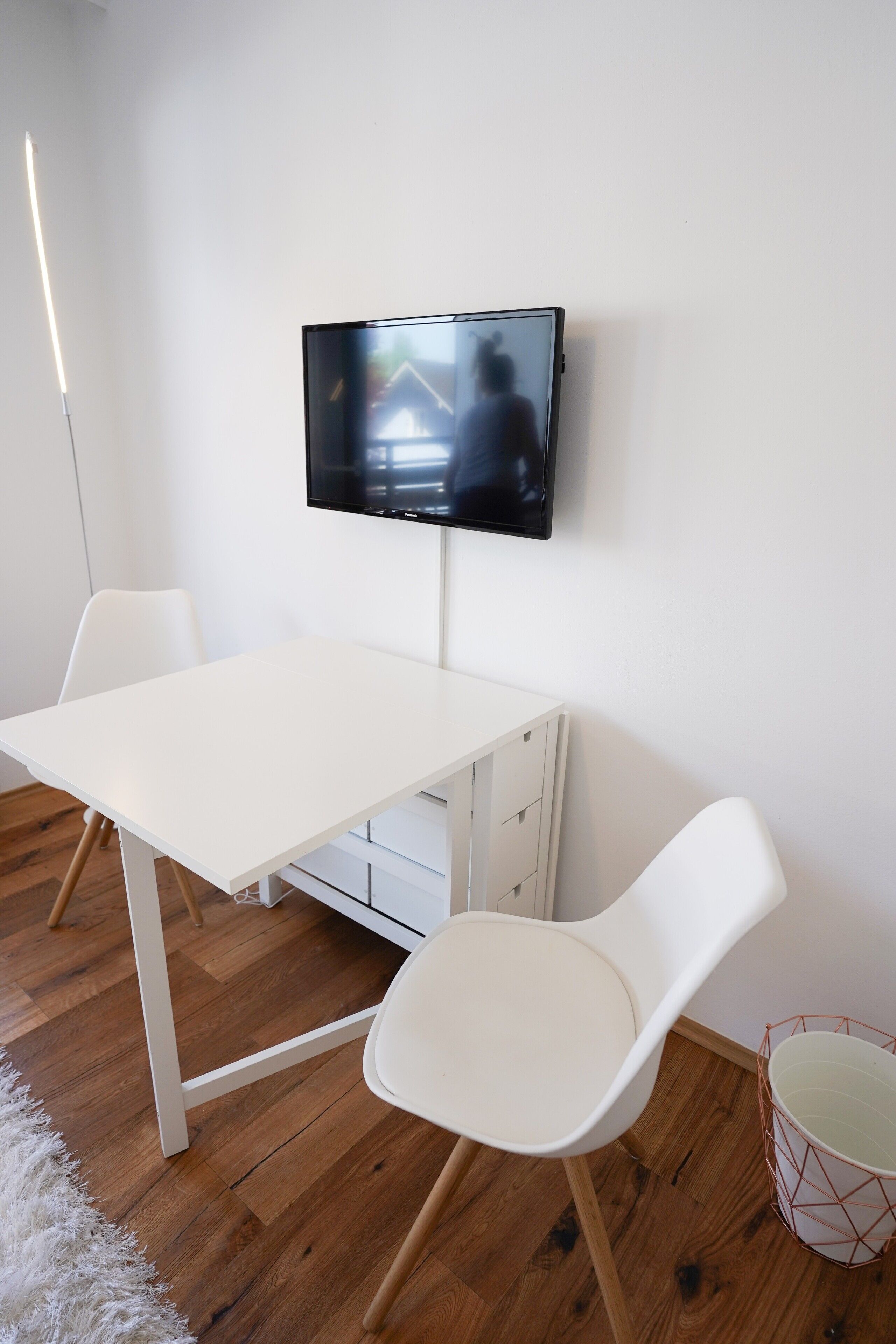Una stanza con un tavolo bianco, due sedie bianche, una TV fissata al muro, un termosifone bianco e un pavimento in legno.