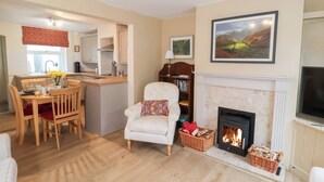 A cozy living room with a fireplace, a dining area with a kitchen in the background, and a bookshelf. - Lilly Cottage (Woodbridge)