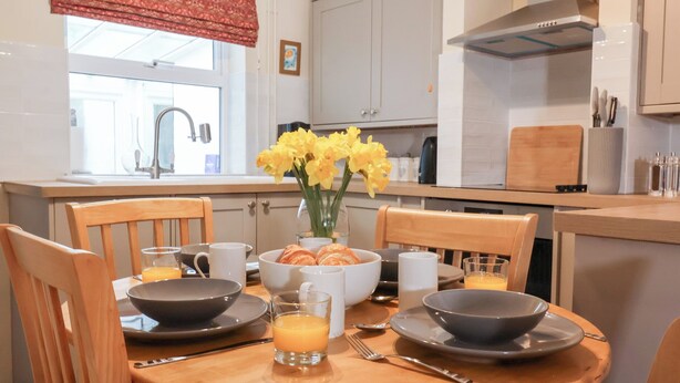 A breakfast setting with a round wooden table, chairs, and a vase of yellow flowers. - Lilly Cottage (Woodbridge)