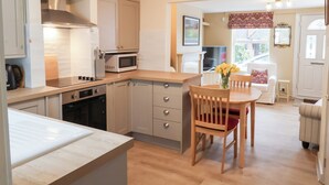A compact kitchen with a microwave, stove, and cabinets. There is a dining area with a table and chairs, and a window with a view of the outside. - Lilly Cottage (Woodbridge)