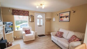 A cozy living room with a white sofa, a white armchair, and a wooden table. There is a television on a stand, a window with a view of the outside, and a door with a mirror above it. - Lilly Cottage (Woodbridge)