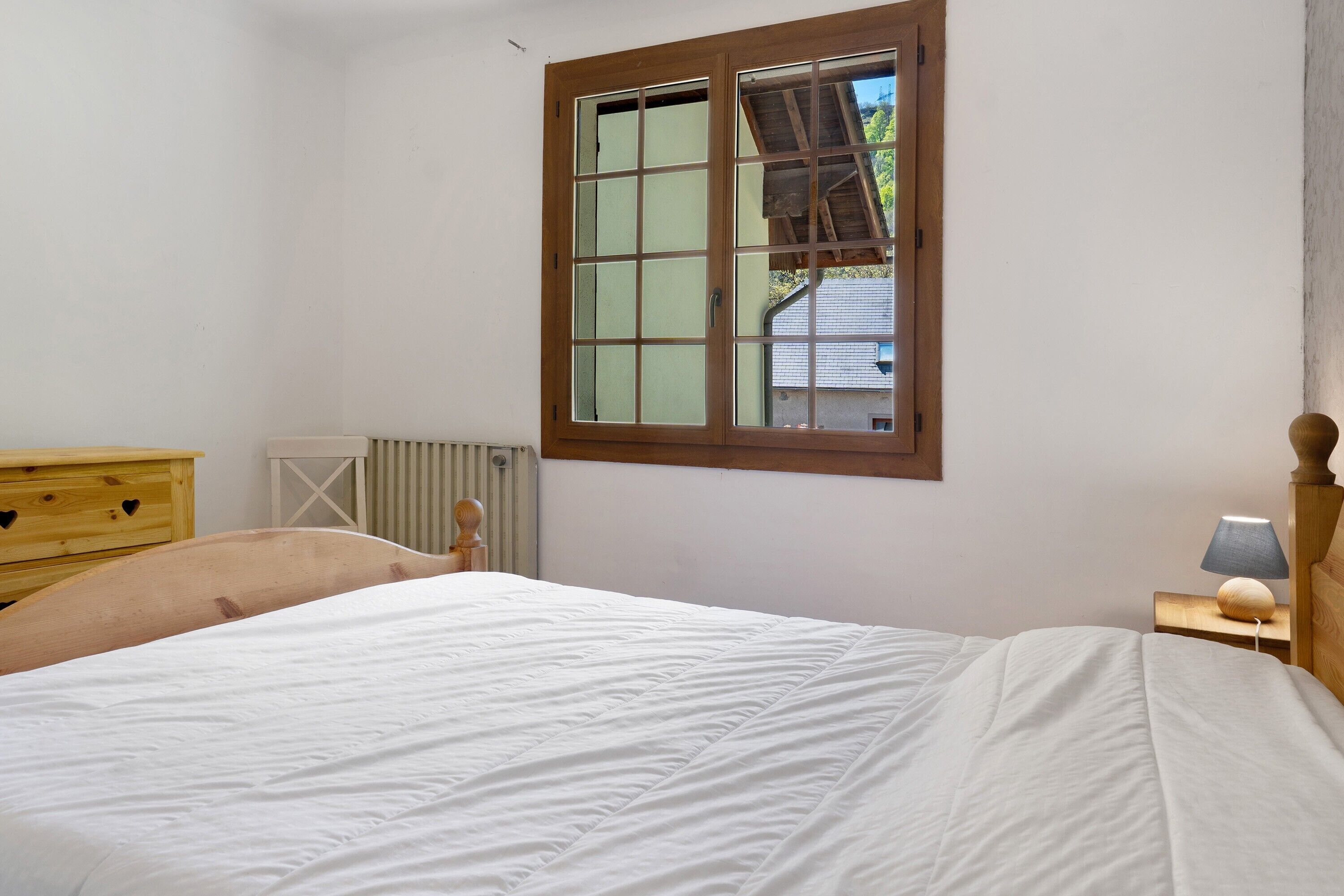 A bedroom with a bed, a wooden dresser, a window, and a bedside table with a lamp.