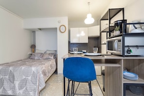 Quarto compacto com uma cama, uma mesa de jantar com cadeiras, uma cozinha compacta com micro-ondas e uma pequena prateleira com utensílios de cozinha.