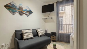Ein modernes Wohnzimmer mit einer Couch, einem kleinen Tisch, einem an der Wand befestigten Fernseher und einem Balkon mit einem gemusterten Vorhang.