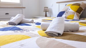 A neatly made bed with rolled towels, a quilt with geometric patterns, a bedside table with a lamp, and a window with daylight.