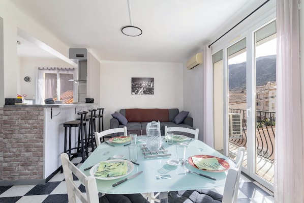 Un coin repas avec une table dressée pour deux personnes, une cuisine en arrière-plan et un balcon avec vue.
