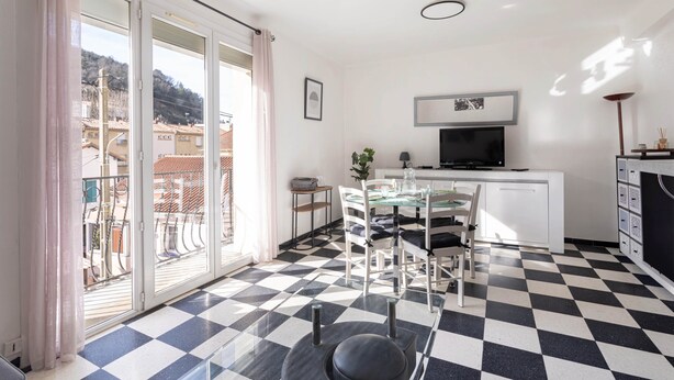 A room with a checkered floor, a dining table with chairs, a TV on a white cabinet, and a balcony with a view of buildings.