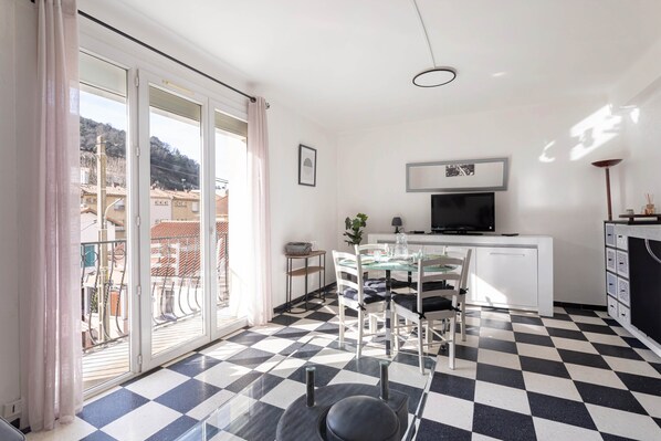 A room with a checkered floor, a dining table with chairs, a TV on a white cabinet, and a balcony with a view of buildings.