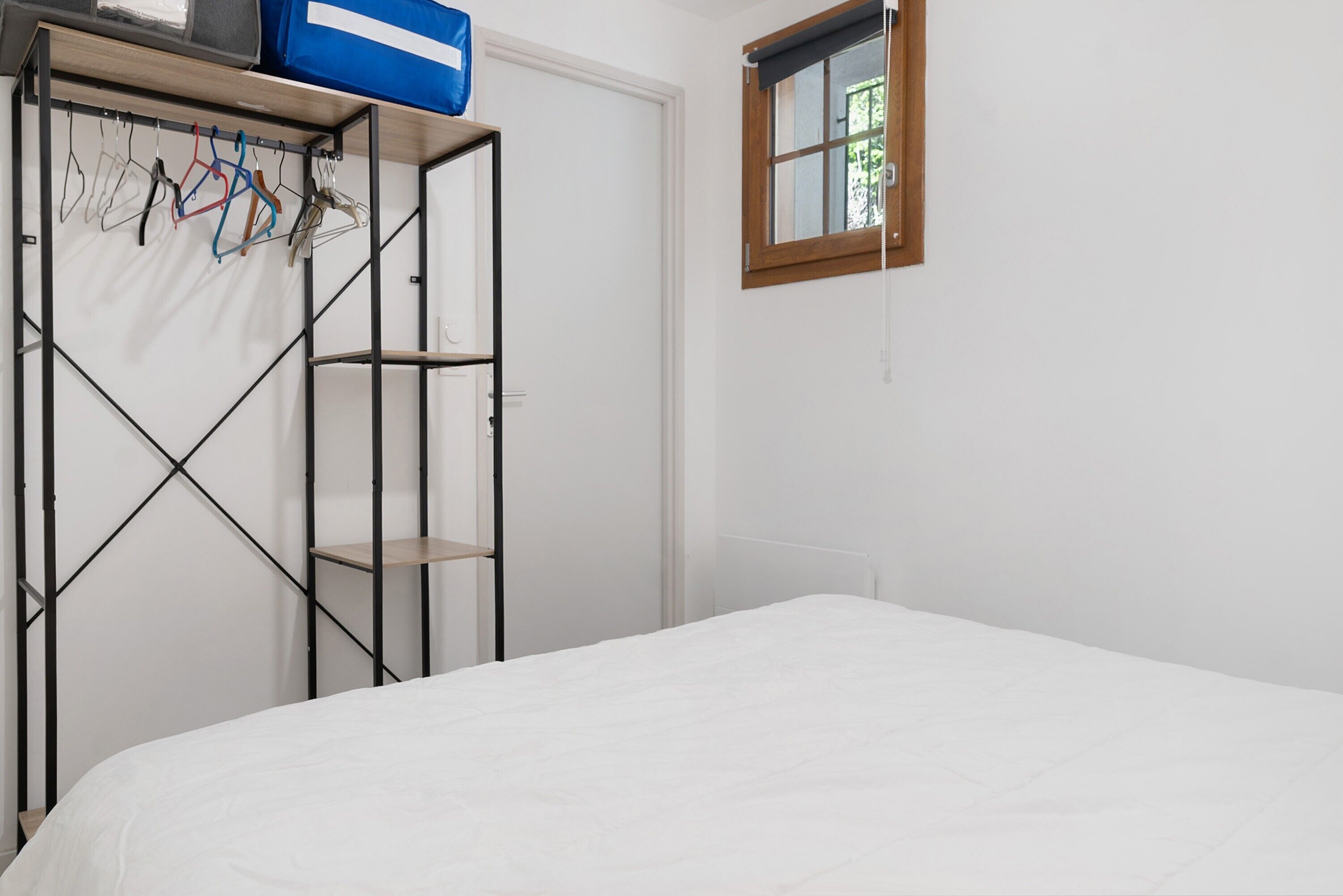 A bedroom with a bed, a wardrobe with hanging clothes, and a window with a view.
