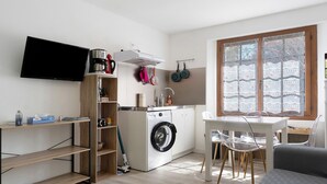 A compact living space with a washing machine, a dining table with chairs, a TV mounted on the wall, and a window with a view. - Apartment 'L'arrieulat 2' with Mountain View, Shared Terrace and Shared Garden (Argelès-Gazost)