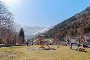 Un parque infantil con tobogán y estructura de escalada, mesas de picnic y casas al fondo.
