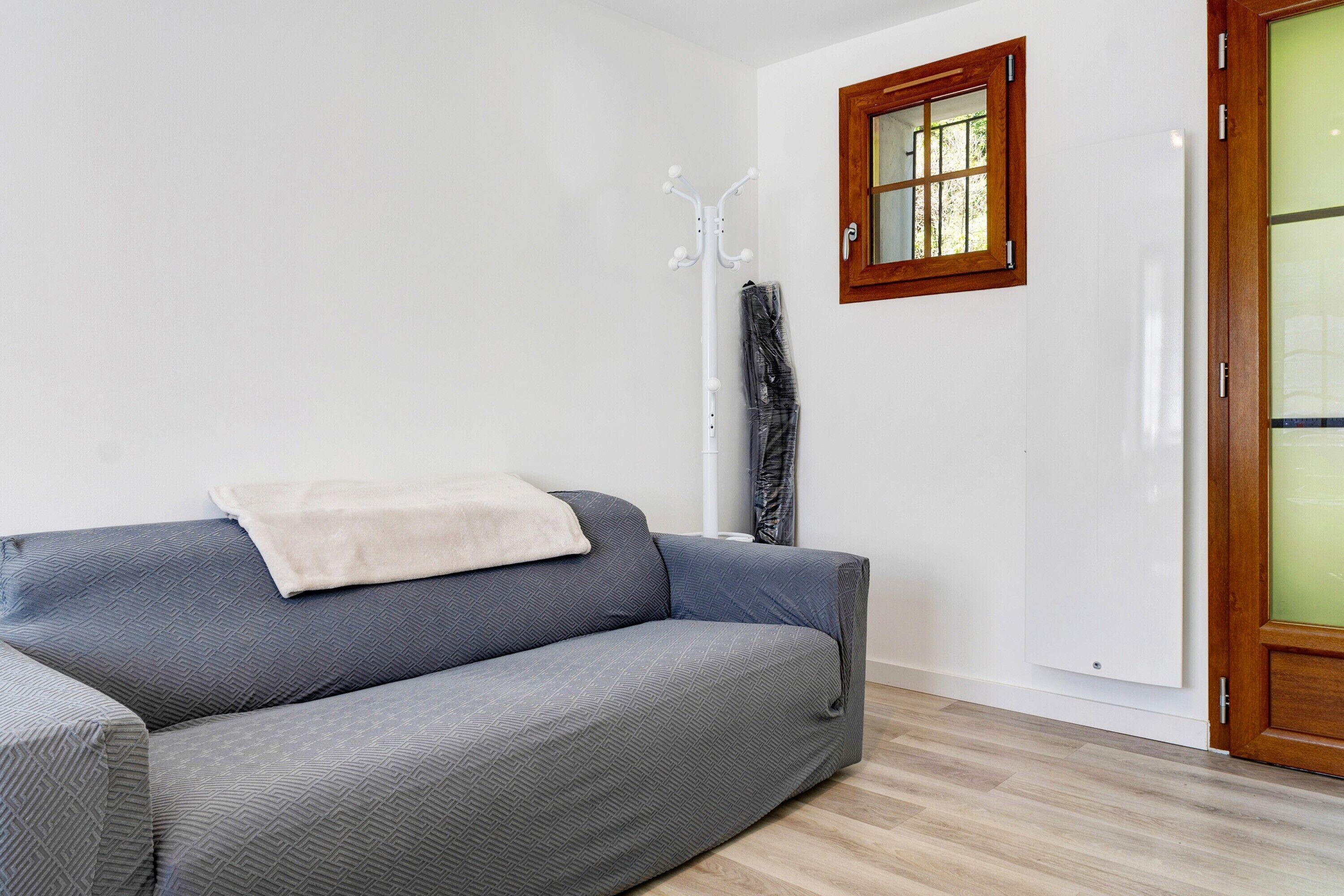 En grå soffa med en vit pläd, en trädörr med en panel av frostat glas och en vit vägg med ett fönster.
