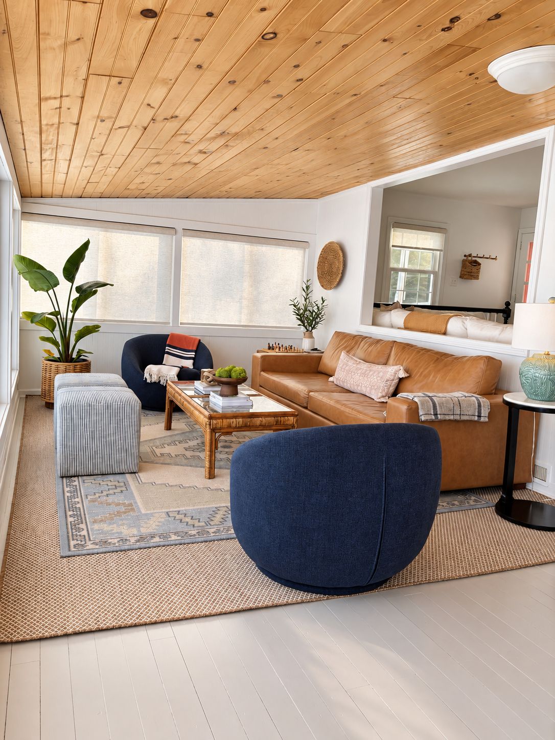 A living room with a wooden ceiling, a blue armchair, a brown sofa, a coffee table with books, and a large window.