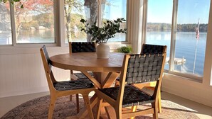 Een eetruimte met een ronde houten tafel, vier stoelen, een potplant en grote ramen met uitzicht op een waterpartij.