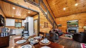 Interior de una cabaña de madera con comedor, cocina, sala de estar, chimenea y una escalera.