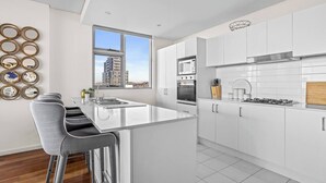 A modern kitchen with white cabinetry, a central island, stainless steel appliances, and a city view through large windows. - City Perch Escape Sweeping Penthouse style Haven (Wollongong)