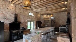 A spacious kitchen with exposed wooden beams, a brick wall, a large island with bar stools, and a built-in oven. - Gîte La Garenne (La Ferté-Saint-Aubin)